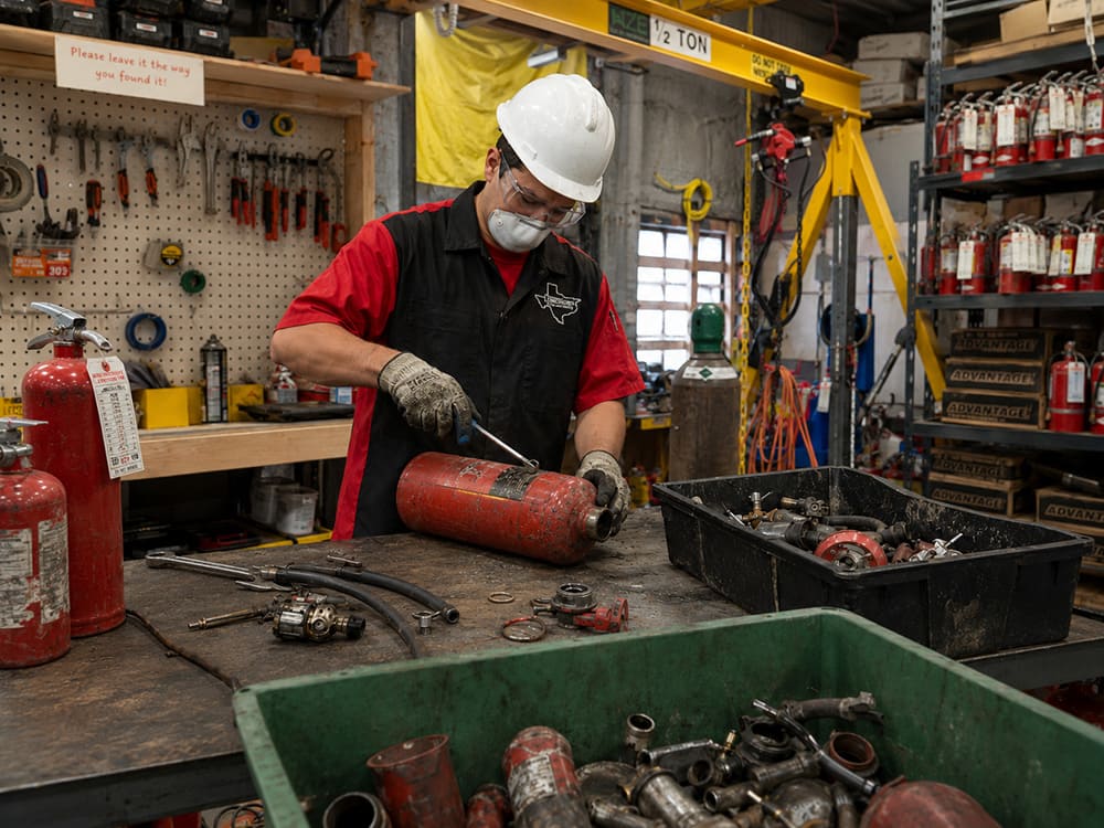 A worker in a hard hat and mask disassembling a red cylindrical device at a cluttered workshop bench, surrounded by tools and parts.